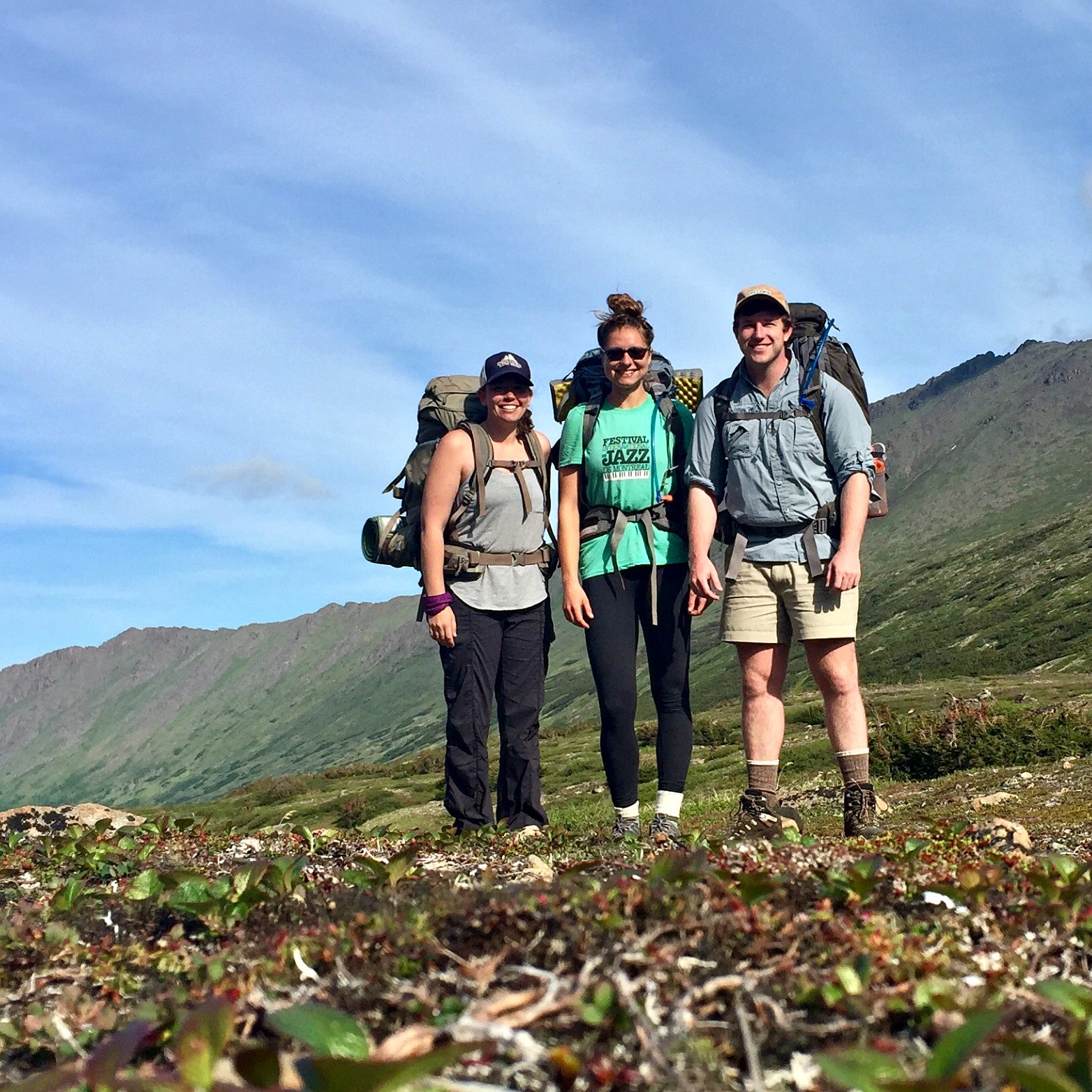 Three hikers stand in front of hills and blue skies in the Chugach Range, Alaska