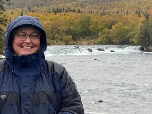 Vicki in cold weather gear with Brooks Falls and fat bears behind her.