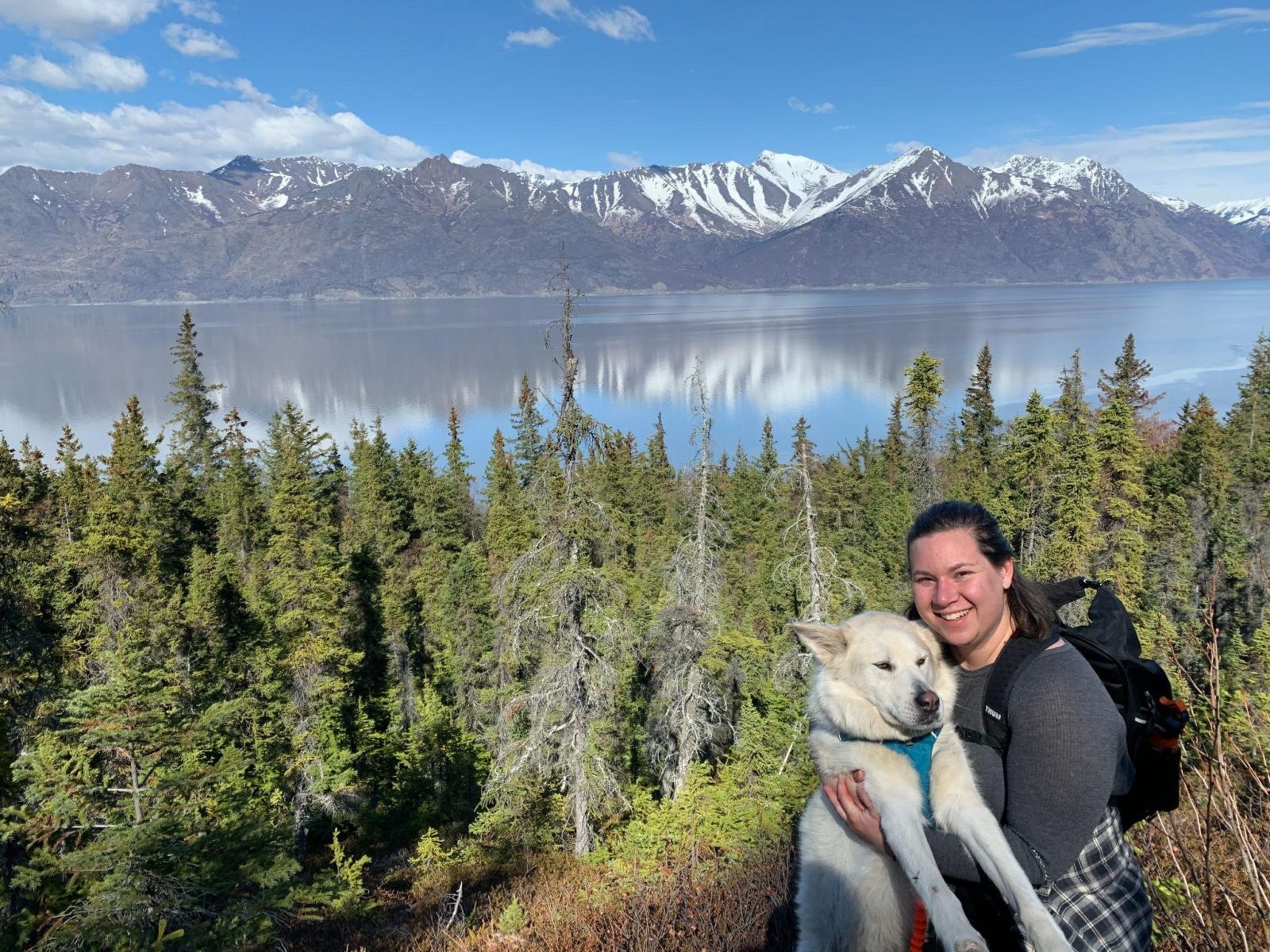 Mackenzie holds a white dog while hiking above a view of the Cook Inlet, reflecting blue sky and mountains 