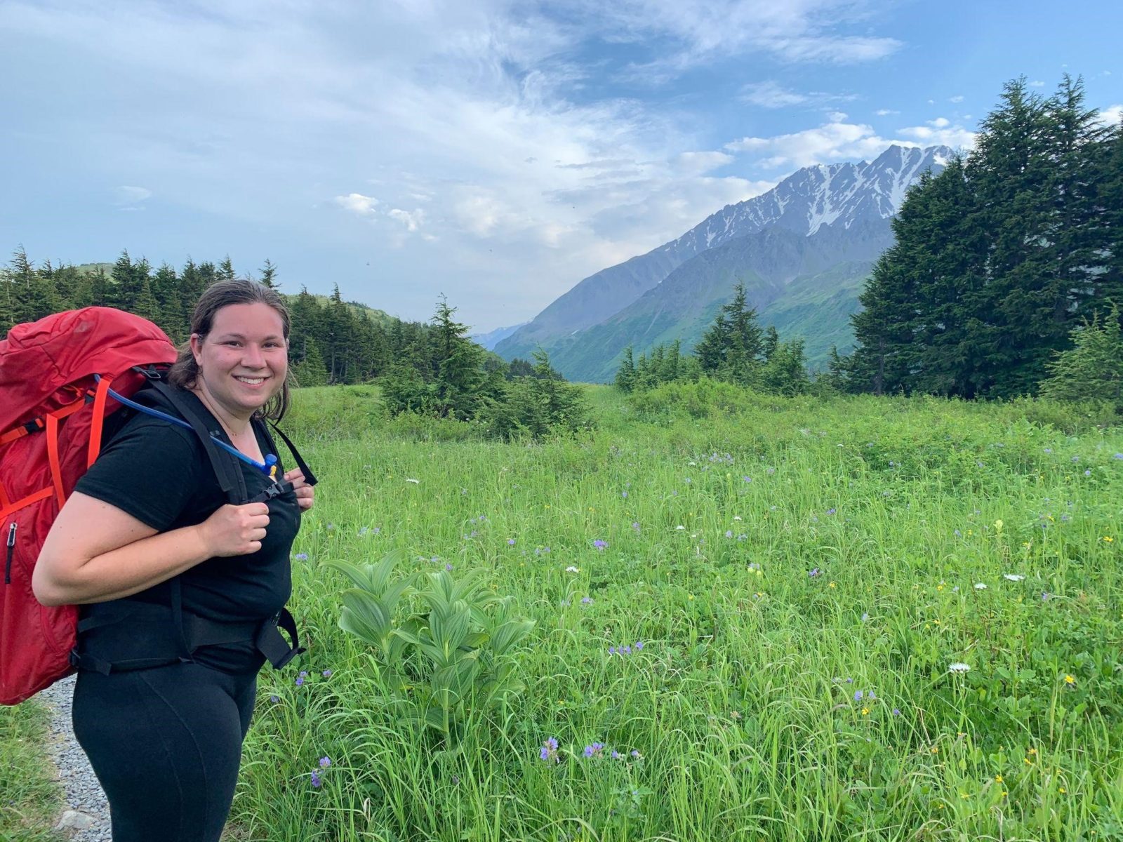 Mackenzie wearing a red backpack in front of a field of wildflowers with a mountain and blue sky while hiking Lost Lake 